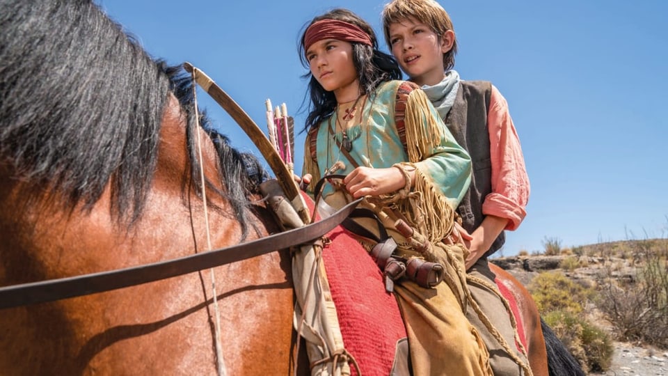 Zwei Jungen auf dem Pferd, vorne mit langen, schwarzen Haaren und Stirnband, hinten dunkelblonder Junge mit Halstuch.