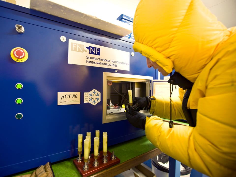 Ein SLF-Mitarbeiter in gelber Daunenjacke und schwarzen Handschuhen stellt etwas in ein grosses, blaues Gerät.