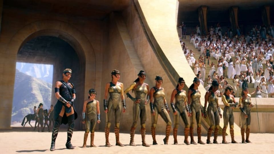 Arena mit hellem Sand. In einer Reihe stehen 7 Frauen in metallenem Outfit, ein Mädchen und eine Frau in schwarz.
