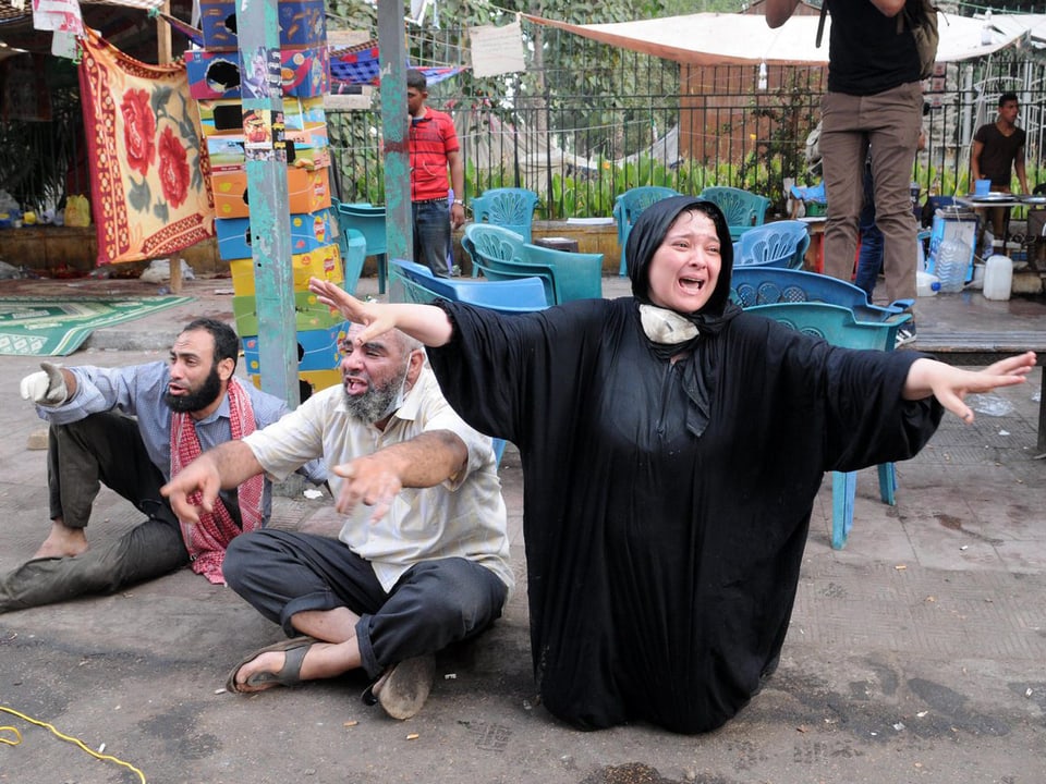 Drei Protestler - davon eine Frau in langem, schwarzem Gewand - knien und sitzen auf dem Boden und halten klagend ihre Hände.
