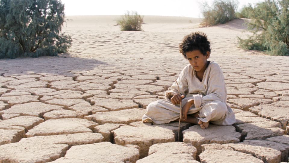 Ein Junge sitzt in einem weissen Gewand auf dem Boden in der Wüste. Der Boden hat Risse.