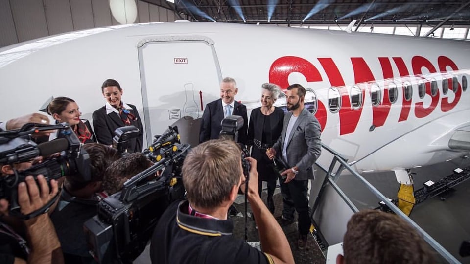 Bligg und zwei weitere Personen vor dem Flugzeug, lächelnd. Mehrere Fotografen und Kameramänner nehmen die drei auf.