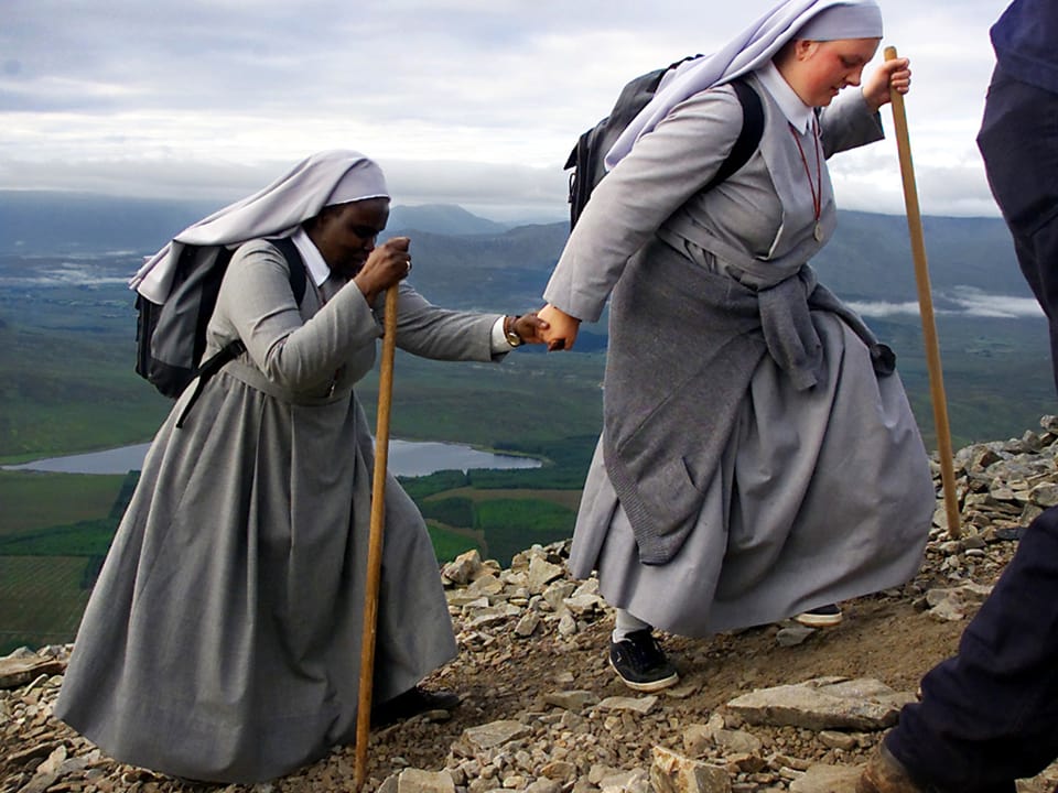 Zwei Nonnen in hellgrauen Gewändern und ausgerüstet mit Holzstock und Turnschuhen gehen Hand-in-Hand auf einem schmalen Pfad.