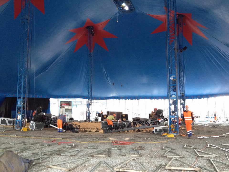 Unter dem blauen Hauptzelt des Zirkus Knie. Metallteile liegen am Boden und Arbeiter montieren die Lichtanlage.