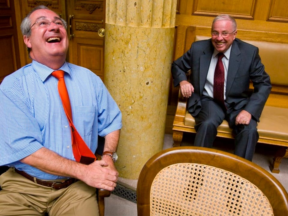 Boris Banga sitzt im Vordergrund und lacht herzlich. Im Hingergrund sitzt Christoph Blocher auf einer Bank und grinst breit. 