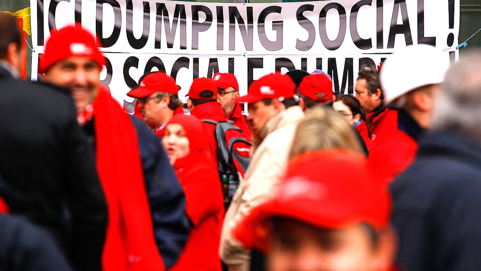 Rot gekleidete Demonstranten in den Strassen Brüssels. Im Hintergrund ist ein Transparent zu sehen mit der Aufschrift «Dumping Social».