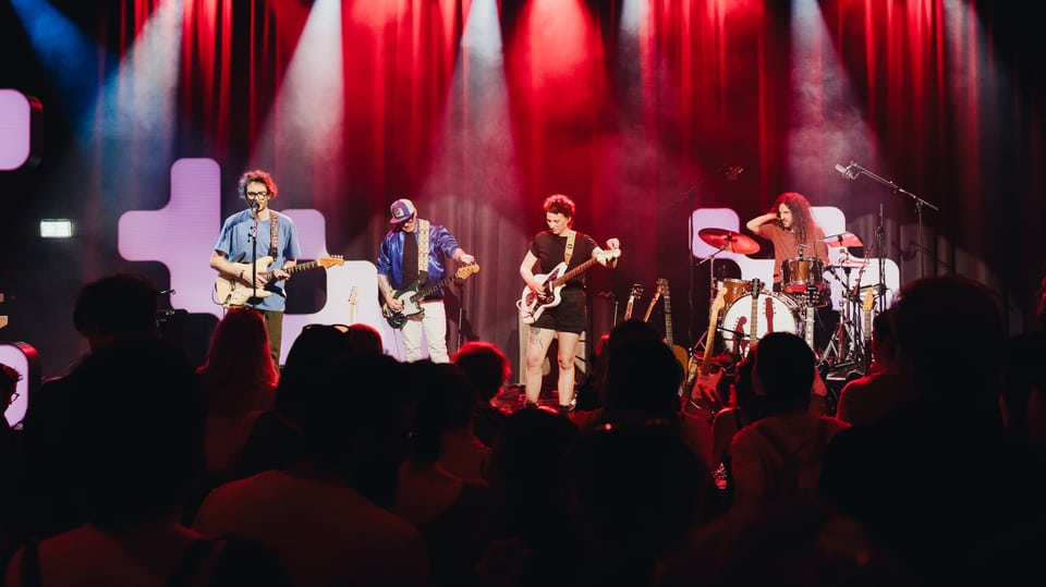 Band spielt live auf einer Bühne vor Publikum bei einem Konzert mit roter Beleuchtung.