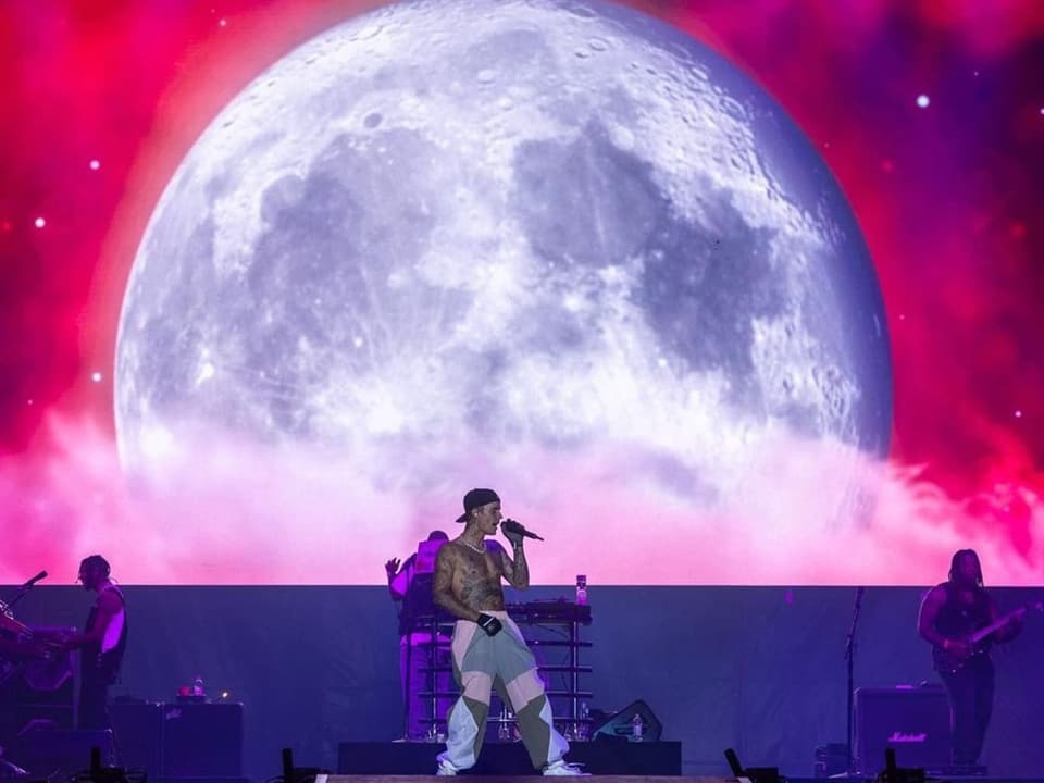 Bild aus der Ferne zeigt den Sänger auf der Bühne. Im Hintergrund ist ein Mondbild mit rotem Himmel.