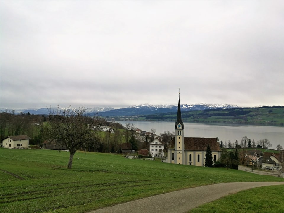 Graue Wolken über der Kirche, im Vordergrund eine grüne Wiese, im Hintergrund der Sempachersee.
