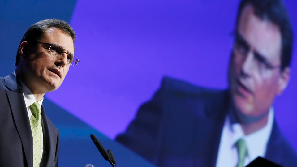 Thomas Jordan hält eine Rede vor blauem Hintergrund, auf dem sein Bild projiziert ist.