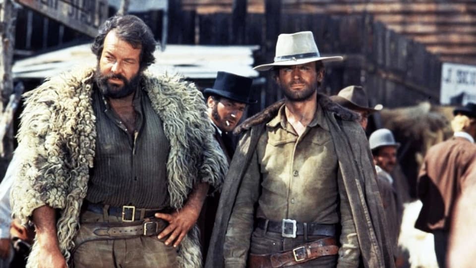 Zwei Männer stehen in einer Western-Stadt. Der eine trägt eine Felljacke, der anderen einen Cowboy-Hut.
