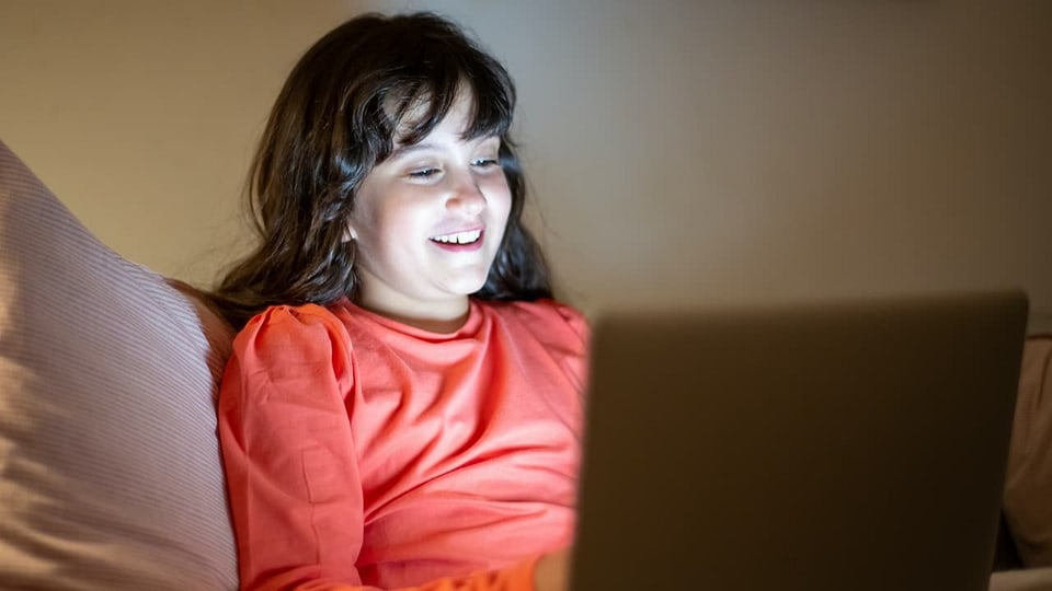 Ein Mädchen sitzt auf dem Bett und liest im Computer.
