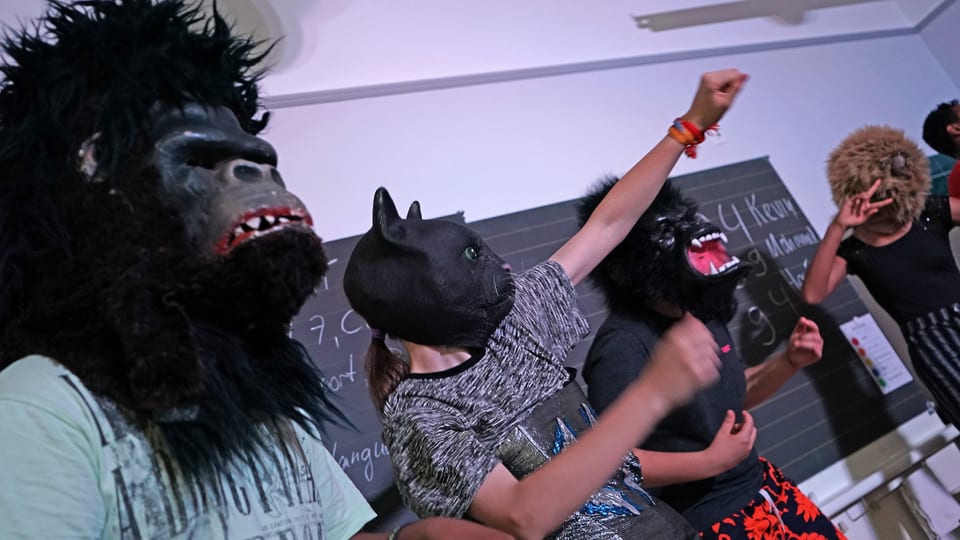 Schüler in T-Shirts vor einer Wandtafel mit schwarzen Masken, schwarzen Haaren, schwarzen Bärten