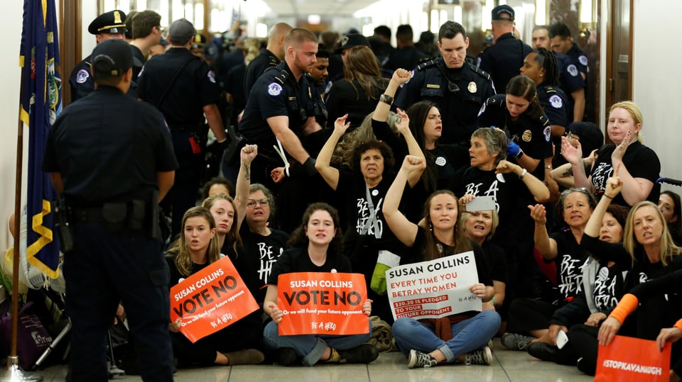 Frauen sitzen in einem Gang und rufen Slogans, sie halten Plakate in den Händen, darum herum stehen Polizisten.
