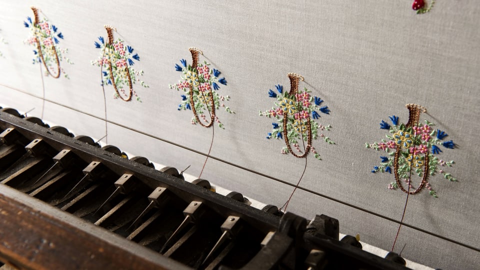 Eine klavierähnliche Tastatur, verbunden mit einer Stoffwand, die Stickereien trägt.