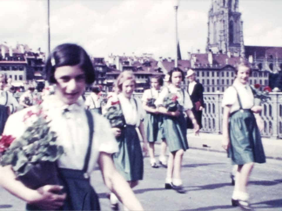 Altes Filmbild: Mädchen marschieren mit Geranien unterm Arm über eine Brücke. 