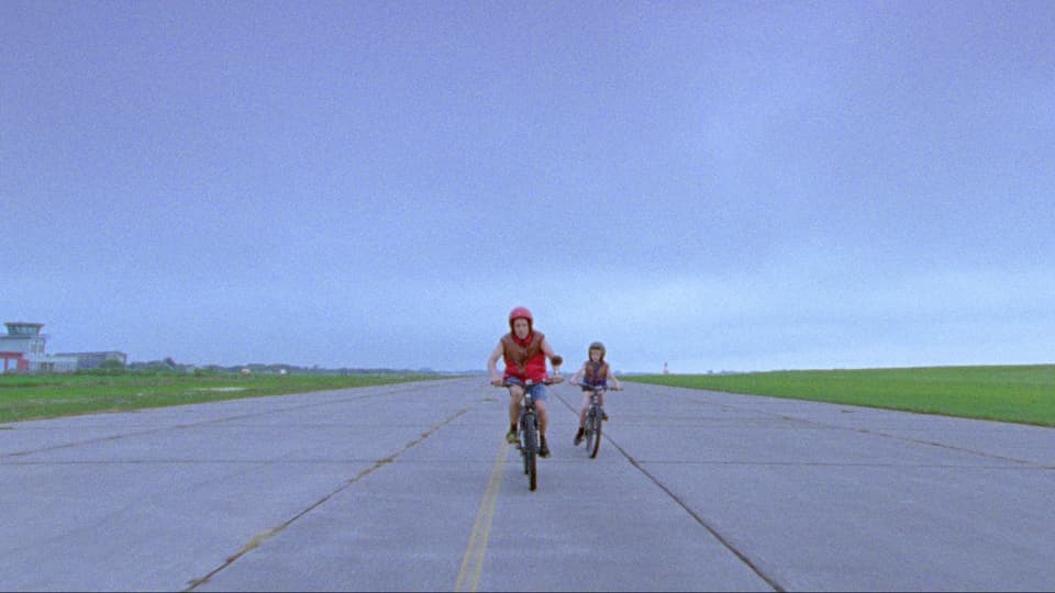 Ein Mann und ein Kind fahren mit dem Velo auf einem abgelegenen Flughafengelände. 