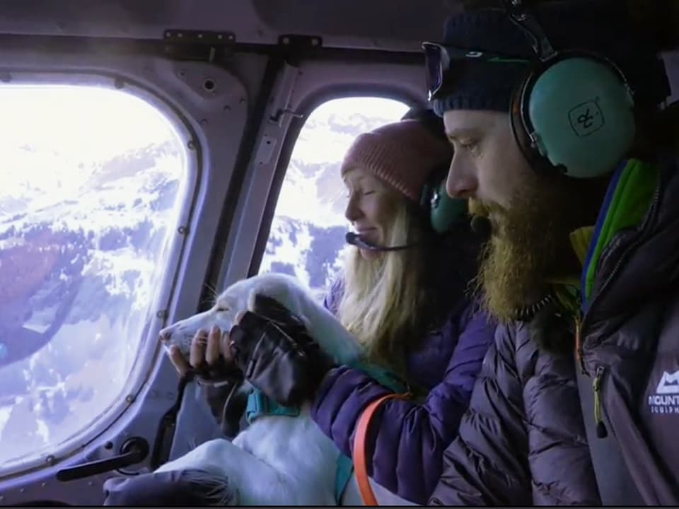 Christina Stewart und Joel Perreten und Hündin schauen aus dem fliegenden Helikopter auf die schneebedeckten Berge.