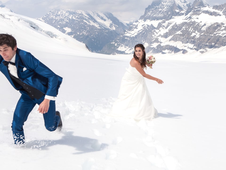 Auf einem Schneebedeckten Gletscher wirft eine Braut im Kleid und mit Blumenstrauss ihrem aus dem Bild flüchtenden Bräutigam einen Schneeball nach.