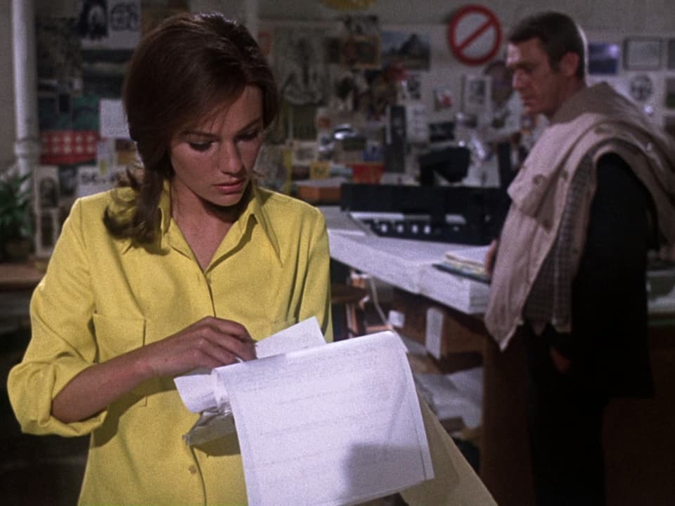 Eine Frau in gelber Bluse liest stehend ein weisses Blatt Papier, hinter ihr im Büro stht ein Mann mit seiner Jacke über der Schulter.