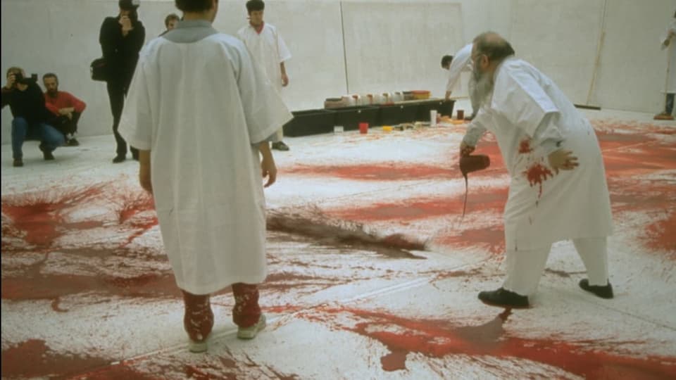 Mann in weisser Kleidung schüttet Blut auf weiss ausgelegten Boden