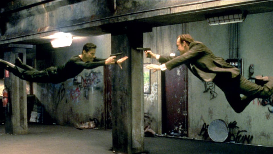 Dois homens voam pelo ar e apontam armas um para o outro.