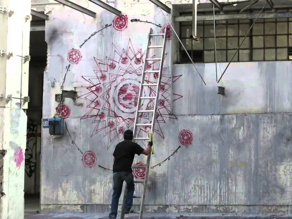 Ein Künstler steigt auf eine Leiter, vor einer mit roten Ornamenten verzierten Betonwand.
