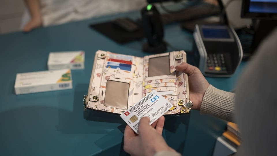 Jemand hält ein Portemonnaie und eine Krankenkassenkarte in der Hand, im Hintergrund eine Zahlmaschine