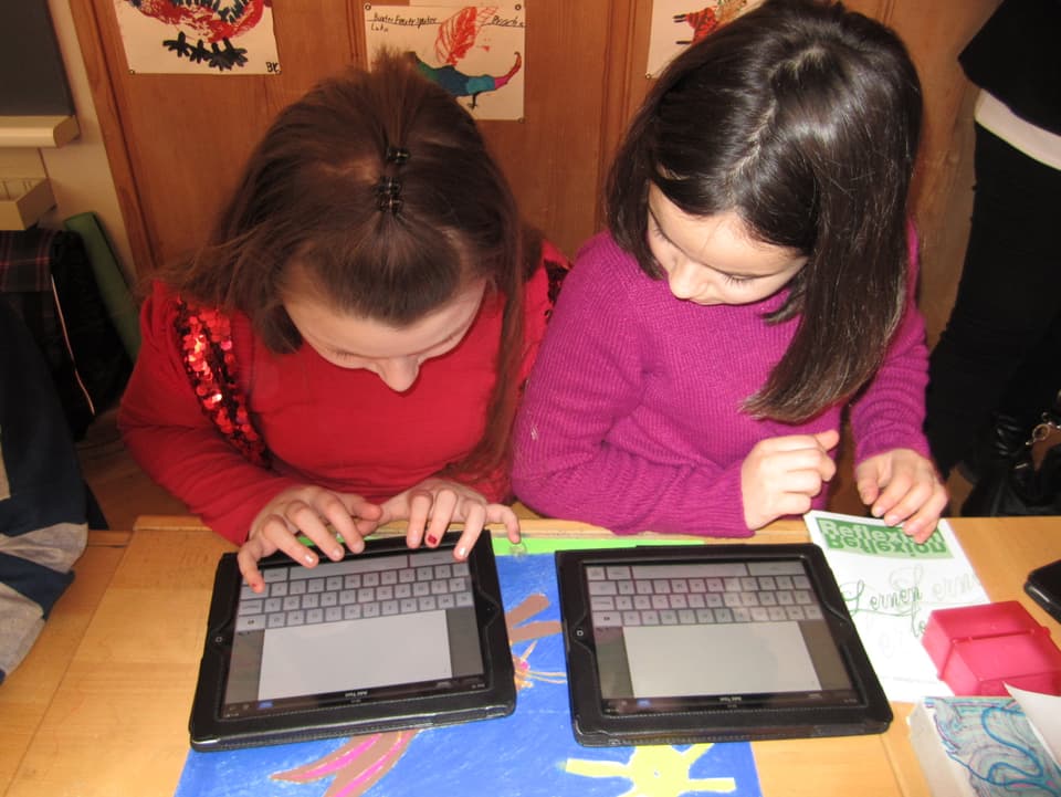 Zwei Mädchen in rotem und rosa Pulli geben im iPad einen Text ein.