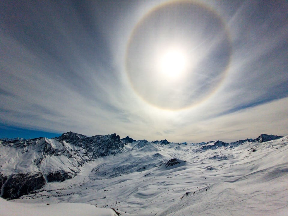 Skigebiet mit Schnee, ziemlich sonnig mit Schleierwolken. In den Wolken ein Kreis um die Sonne. 