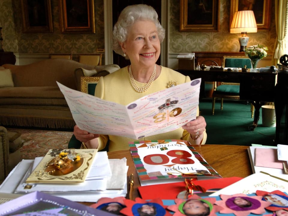 Queen Elizabeth sitzt an einem Tisch und liest eine grosse Karte, die sie zum 80. Geburtstag erhalten hat. Vor ihr liegen weitere Geburtstagskarten.