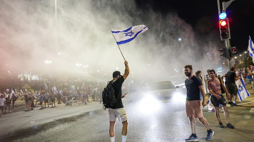 Ein Mann schwenkt die israelische Flagge auf der Strasse; um ihn sind Menschen, Autos und Rauch zu sehen.