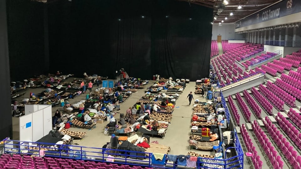 Eine Halle mit vielen Betten am Boden, daneben pinke Stühle einer Tribüne.