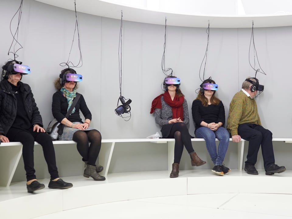 Besucher sitzen auf einer Bank mit VR-Brillen auf dem Kopf.