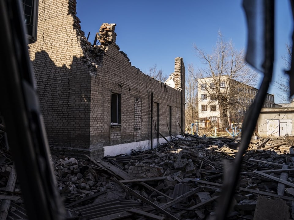 zerstörtes Gebäude, Dach fehlt, davor am Boden Schutt, im Hintergrund Wohnblock