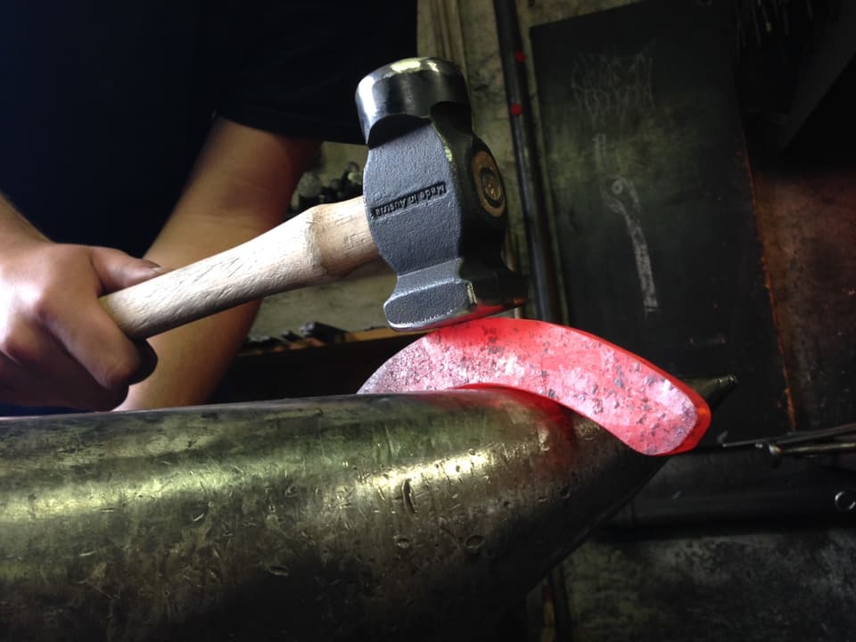 Hufschmied schlägt mit Hammer auf glühendes Eisen, das über einem runden Rohr liegt.