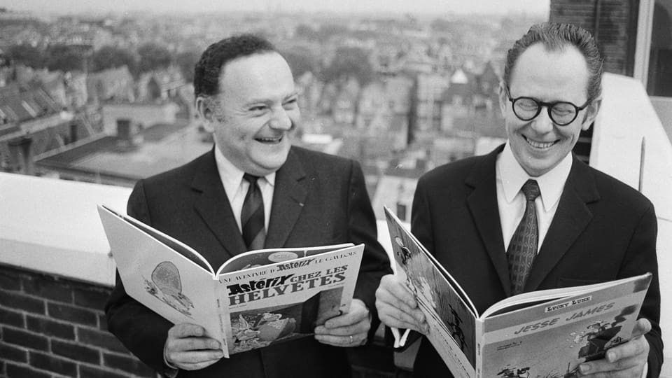 Zwei alte Männer in Anzug halten Comicbücher in der Hand. Sie lachen.