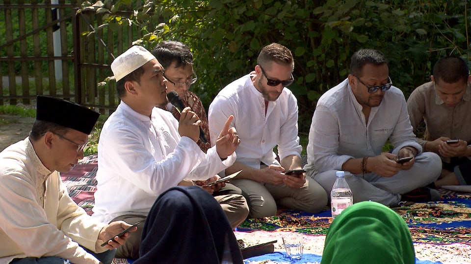 Männer sitzen auf Teppichen rund um einen Imam, der in ein Mikrofon spricht