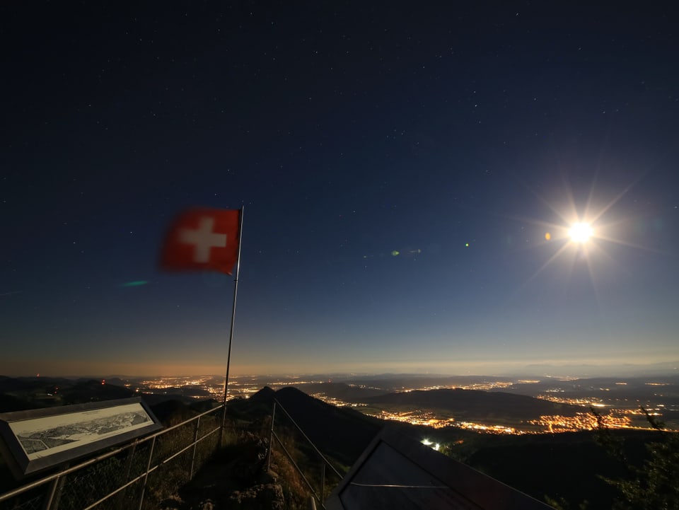 Der Vollmond erhellt die Nacht. Im Mittelland sind die Lichter der Städte zu sehen. Die Schweizer Fahne weht im Wind.