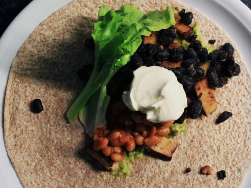 Tortilla-Fladen mit Salat, Crème fraîche, Bohnen und Tofu auf einem Teller.