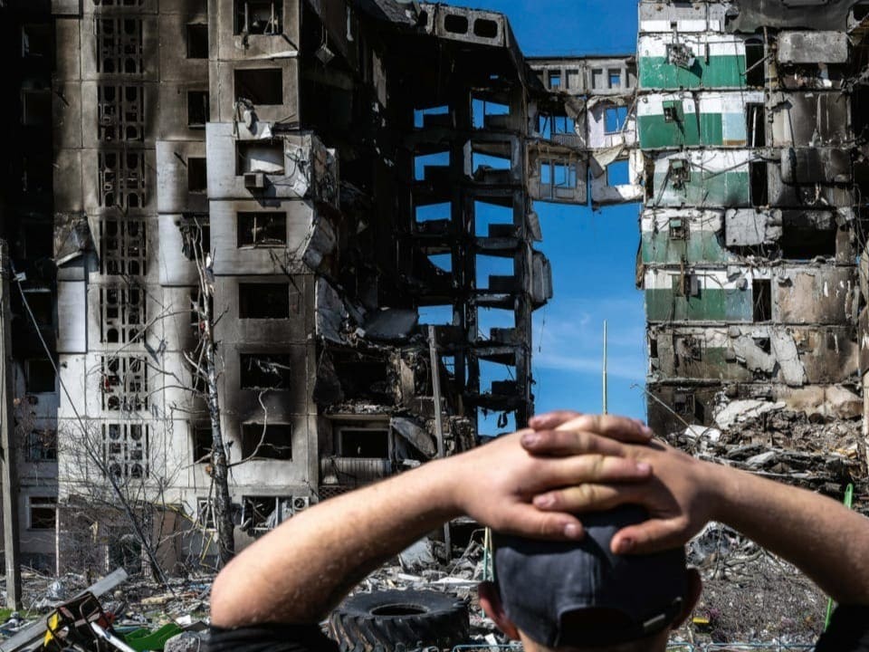 Ein junger Mann verschränkt seine Arme hinter seinem Kopf. Neben ihm ein Mann. Er schaut auf ein bombardiertes Haus.