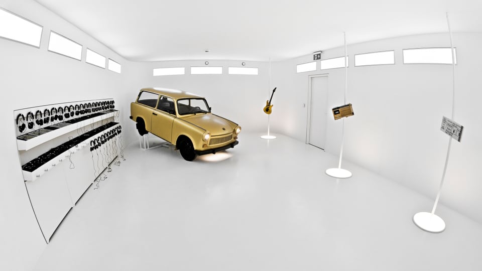 Ein weisser Museumsraum: darin scheint ein gelbes Auto zu schwebe. Daneben hängen viele Kopfhörer.