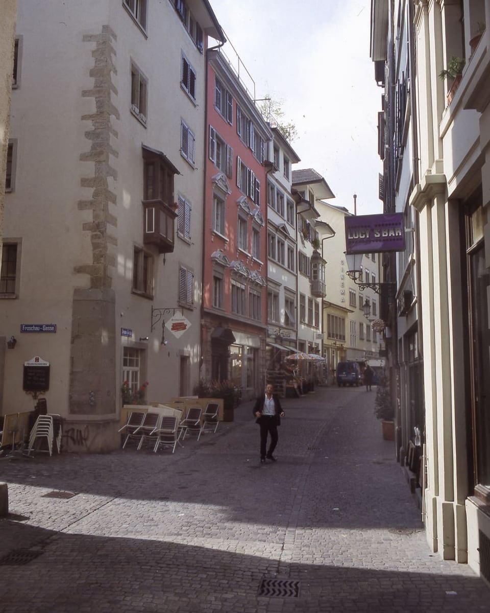 Foto einer Gasse, Pflasterstein, links weisses dann rosa Haus, Stühle links auf der Gasse.