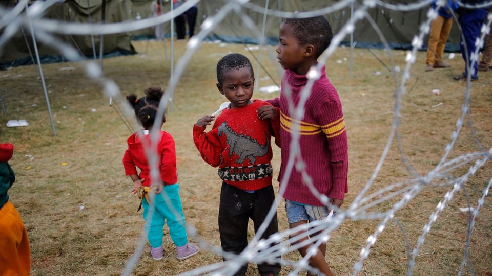 Kinder hinter Stacheldrahtzaun, im Hintergrund Zelte.
