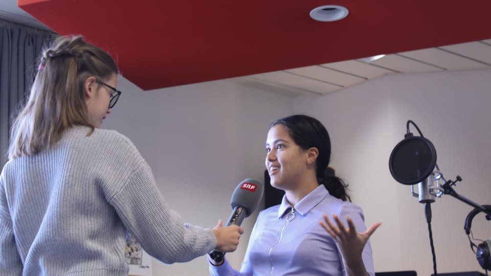 Cachita steht vor Emelie und wird von ihr mit einem Mikrofon interviewt. Im Hintergrund sieht man das Tonstudio.