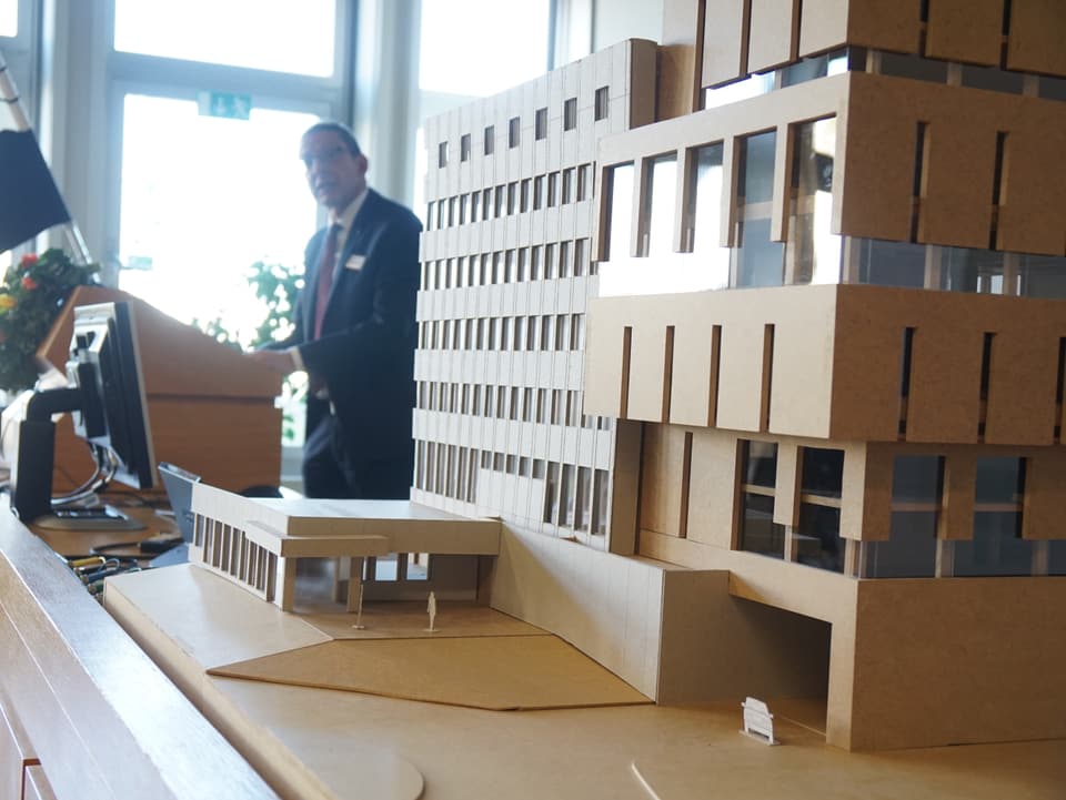 Ein Baumodell des neuen Gebäudes für die Notrufzentrale ist aufgestellt - im Hintergrund sieht man einen Mann, der an einem Rednerpult steht.