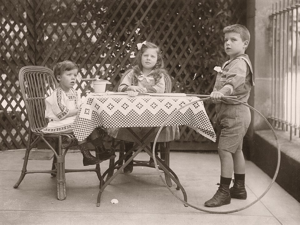 Eine alte Fotografie zeigt drei elegant gekleidete Kinder an einem Tisch.
