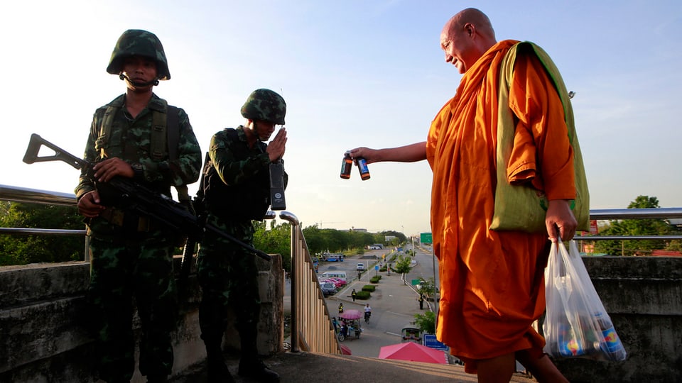 Ein buddhistischer Mönch in rotem Gewand reicht zwei Soldaten ein Getränk.