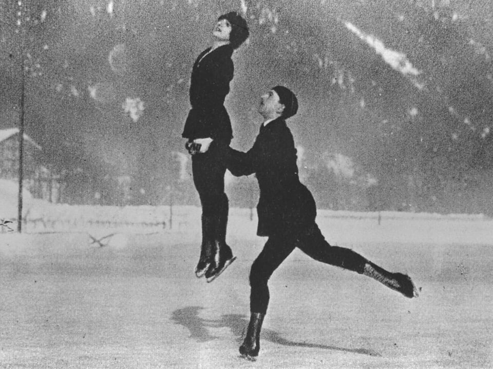 Im Eiskunst-Paarlauf wirft gerade der Mann die Frau in die Luft, beide sind schwarz gekleidet.