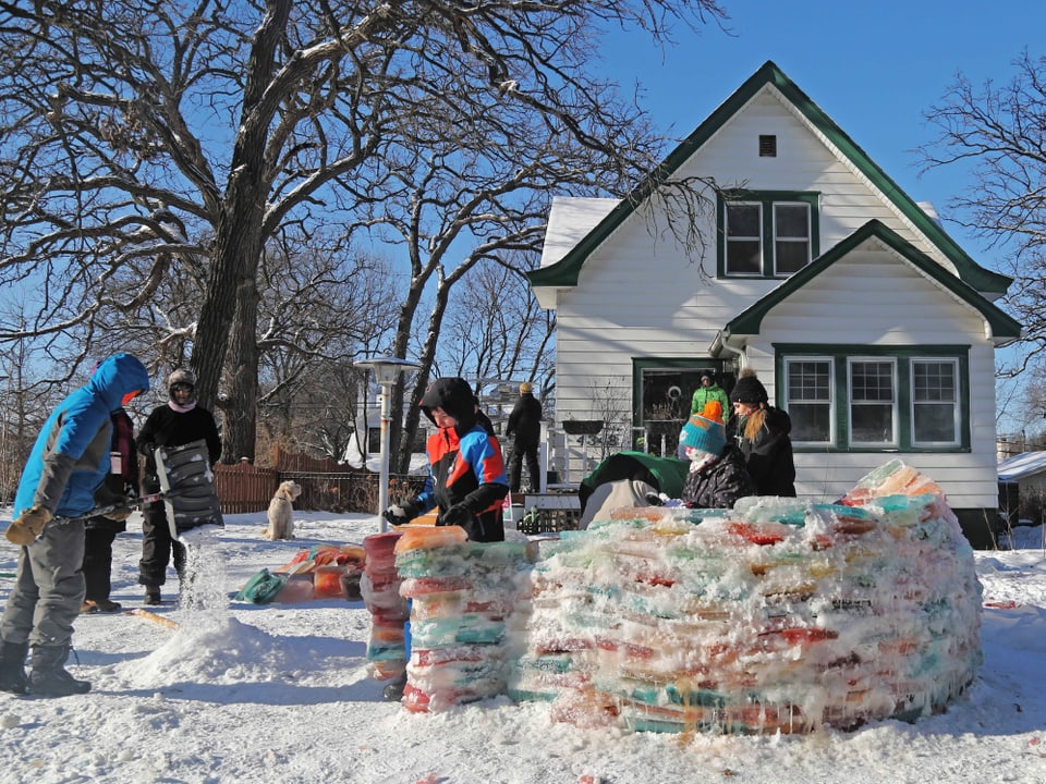 Leute bauen ein buntes Iglu, im Hintergrund ein Haus, Schnee